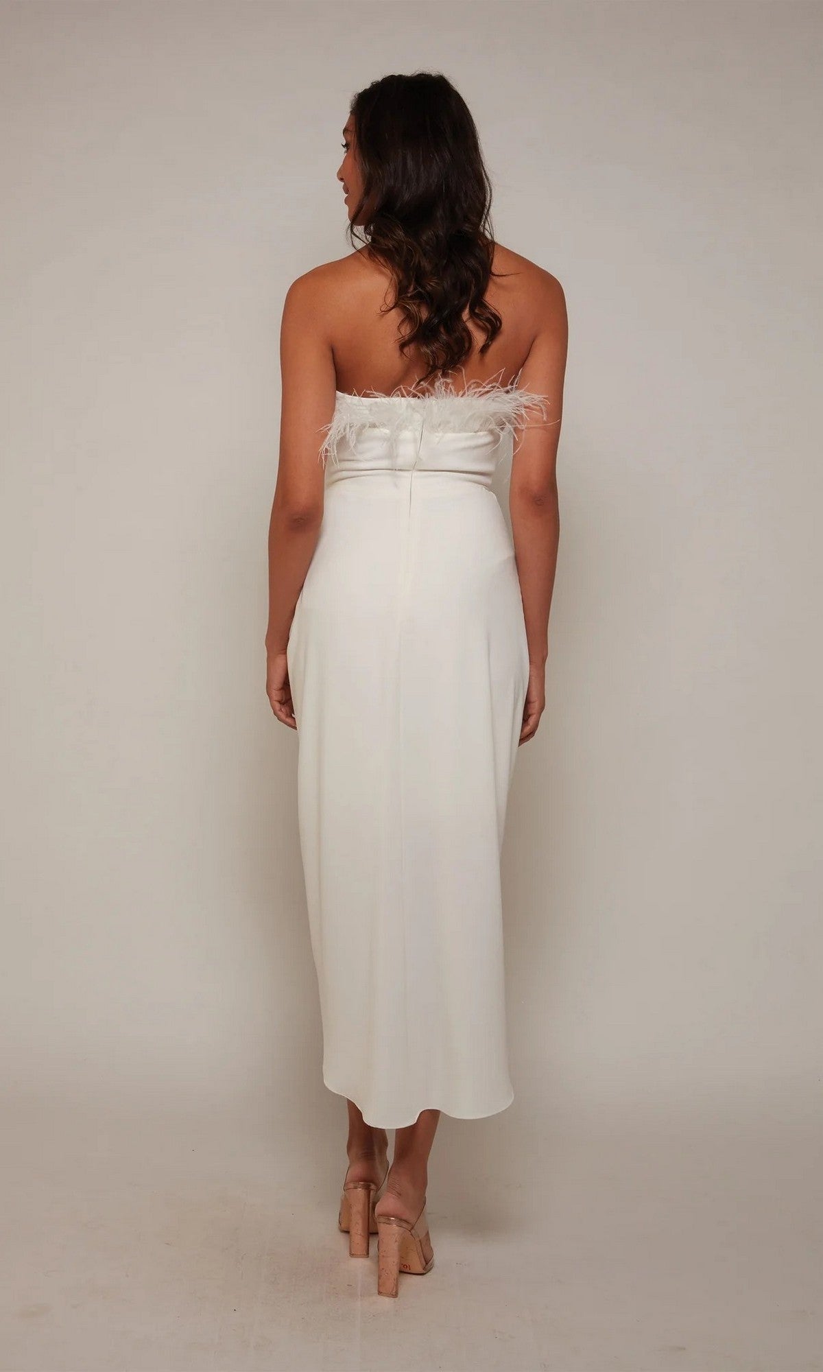 Woman facing away from the camera in a white, strapless Alyce 70034 knee-length semi-formal dress with feather accents, set against a plain background.